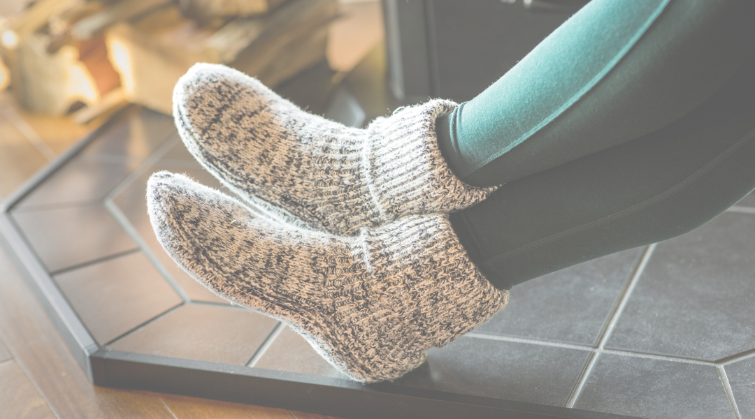 person with feet by fire wering merino wool slippers that are grey and black.  feet are resting on black tile 