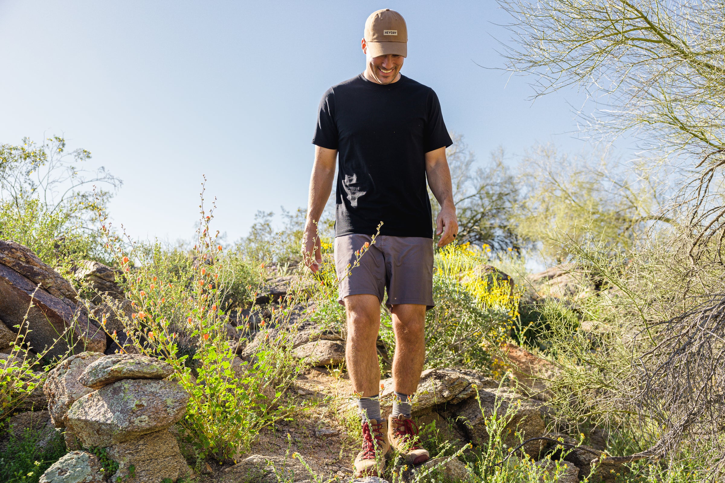 A person wearing a cap, black shirt, shorts, and hiking boots walks down a rocky, desert trail surrounded by plants and clear blue sky.
