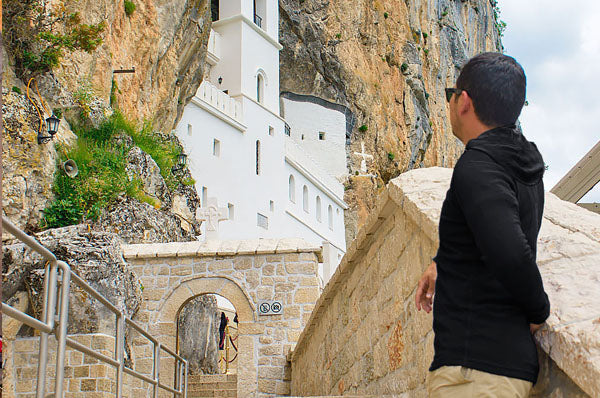 A man in a black hoodie stands on stone steps, looking up at the white Ostrog Monastery built into a rocky cliffside surrounded by natural rock formations.