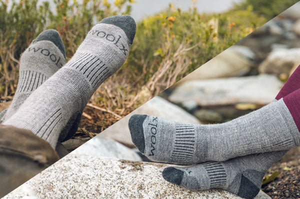 Two pairs of feet wearing gray Woolx socks rest outdoors on rocks, surrounded by greenery, suggesting a comfortable and relaxing moment in nature.