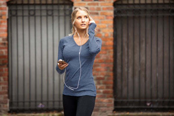 A woman in a blue long-sleeve shirt and black pants stands outside in front of a brick wall, holding a phone, with earphones in her ears, looking to the side.