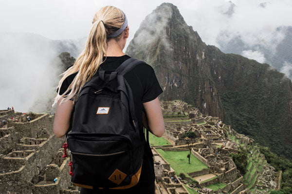 A person with a black backpack stands overlooking the ancient ruins of Machu Picchu, shrouded in mist and surrounded by lush green mountains.