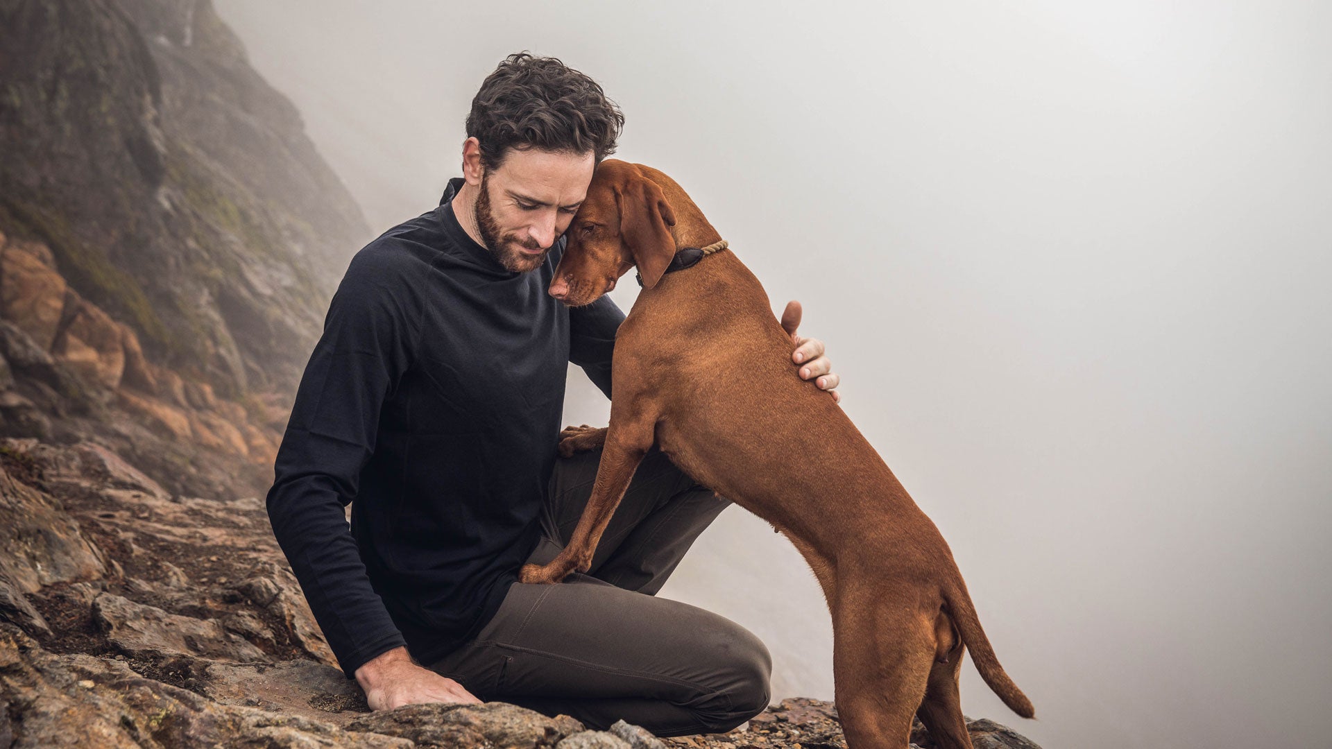 A man kneels on rocky terrain, embracing a brown dog that leans against him. Foggy landscape in the background creates a serene, contemplative atmosphere.