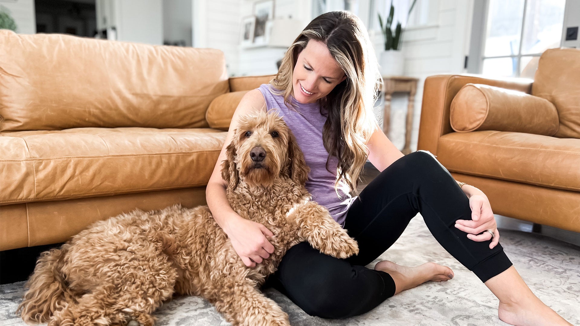 A woman sits on the floor, smiling and petting a fluffy brown dog. They are in a cozy living room with tan leather couches and a light rug.