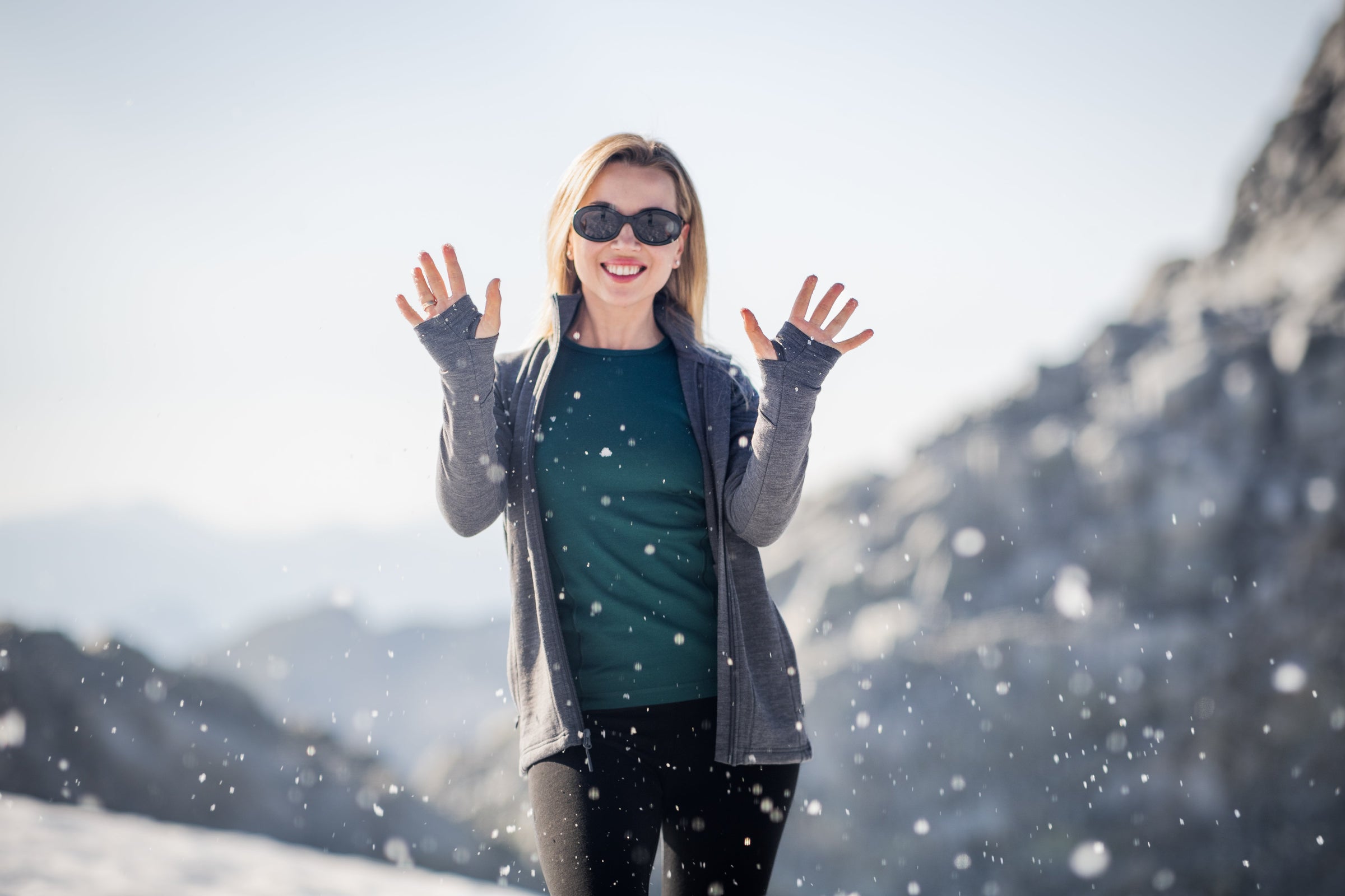 A woman wearing sunglasses and a gray jacket smiles and raises her hands while standing in a snowy landscape with mountains in the background and snow falling around her.