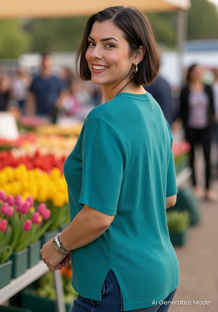 A woman in the Woolx Delia Ribbed Short Sleeve in Surfside smiles by colorful flowers at an outdoor market.