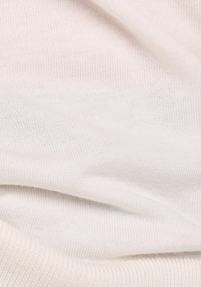 A close-up of the Macie Sweater in Dewdrop Pearl by Woolx shows its soft, cream-colored knitted fabric. The material is slightly folded, creating gentle curves and shadows on its textured surface.