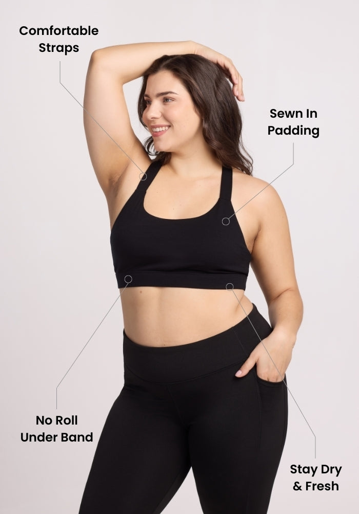 A woman in the Woolx Livi Bra - Black and leggings poses, smiling with one arm raised. Labeled features highlight seamless feel straps, sewn-in padding, a no roll under band, and stay dry & odor-resistant fabric.