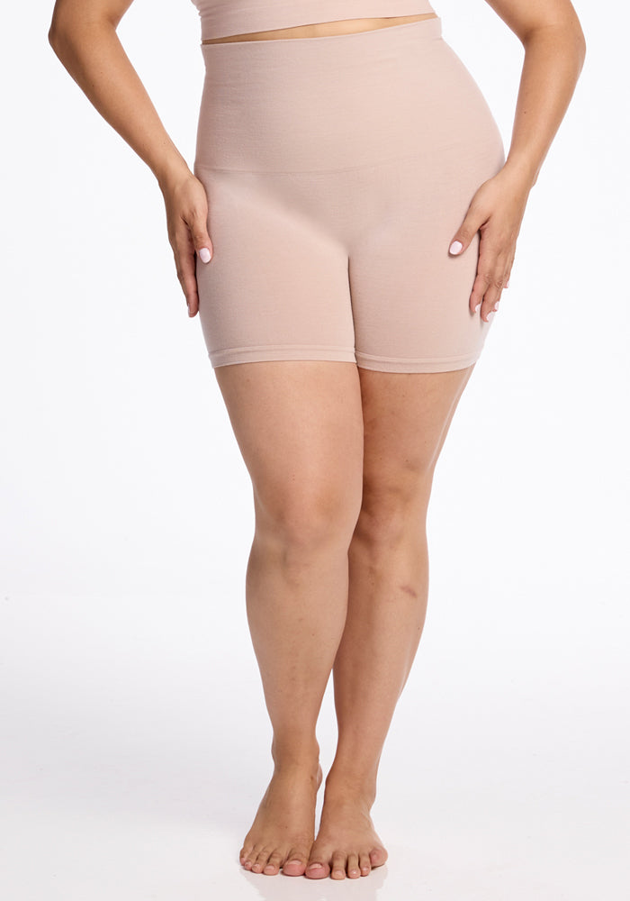 Modeling Woolx's Marley Shapewear Shorts - Sand, a woman stands barefoot against a white background. These high-waisted, seamless shorts extend from her waist to mid-thigh, creating a smooth silhouette as she gently places her hands on her hips.