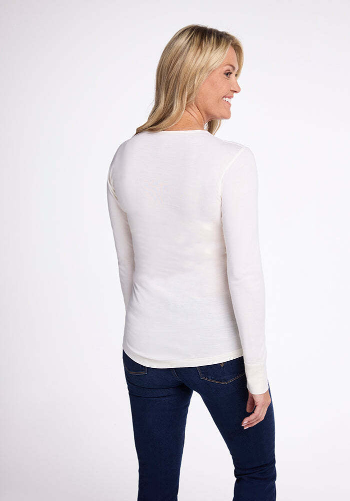 A person with long blonde hair smiles as they stand slightly turned to the side, wearing a moisture-wicking Hannah Long Sleeve Top in Buttercream by Woolx paired with dark jeans, against a plain white background.