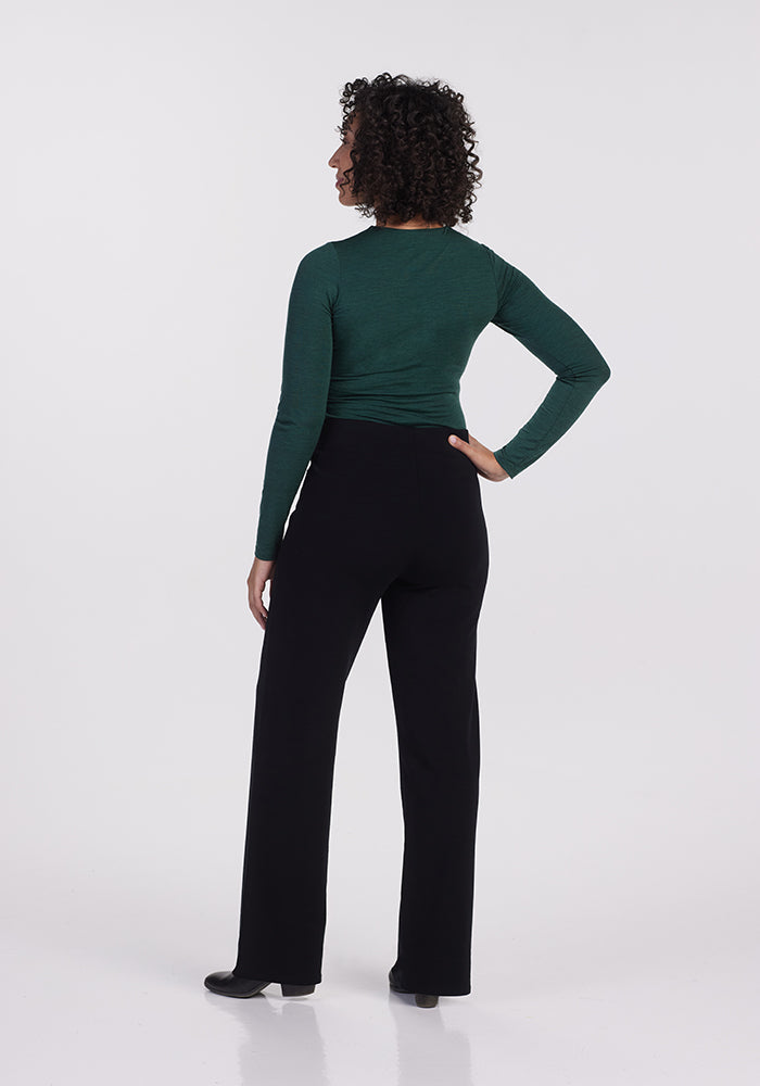 A person with curly hair stands with their back to the camera wearing a long-sleeved green top and the Ellie Wide Leg Pants - Petite from Woolx in black, against a plain white background. They have one hand on their hip.