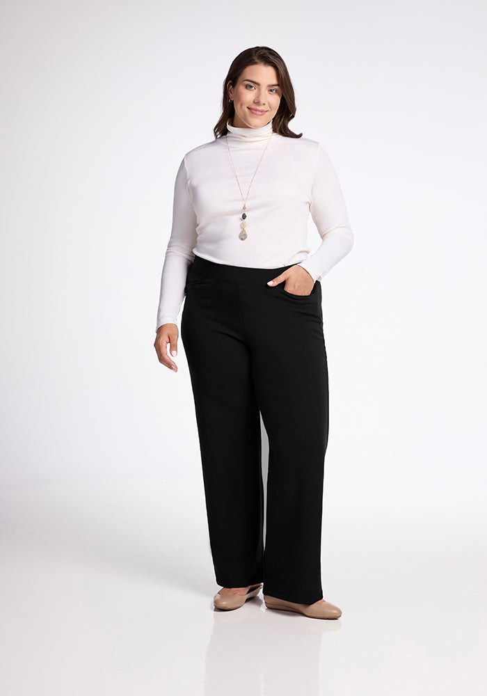A tall woman stands against a plain background, wearing a white turtleneck and the Ellie Wide Leg Pants - Tall by Woolx in black. She pairs her outfit with beige shoes and accessorizes with a long gold necklace featuring a pendant, while keeping her right hand in one of the pants' pockets.