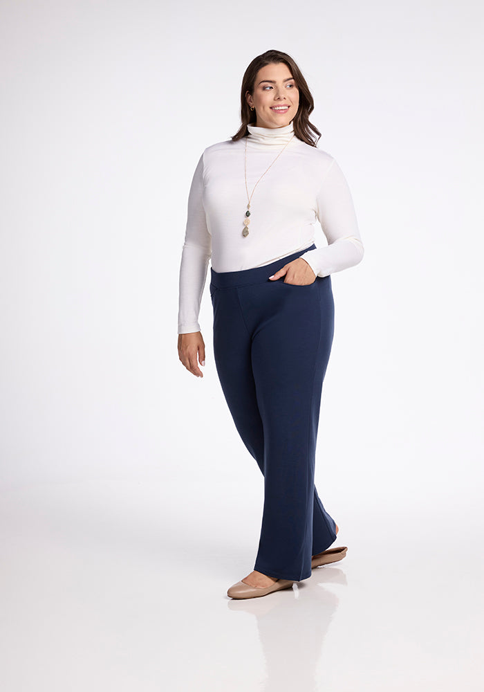 A woman wearing a white turtleneck and the Ellie Wide Leg Pants in Deep Navy from Woolx strikes a confident pose. With her hand in one pocket and a long necklace draped elegantly, she stands against a plain white background.