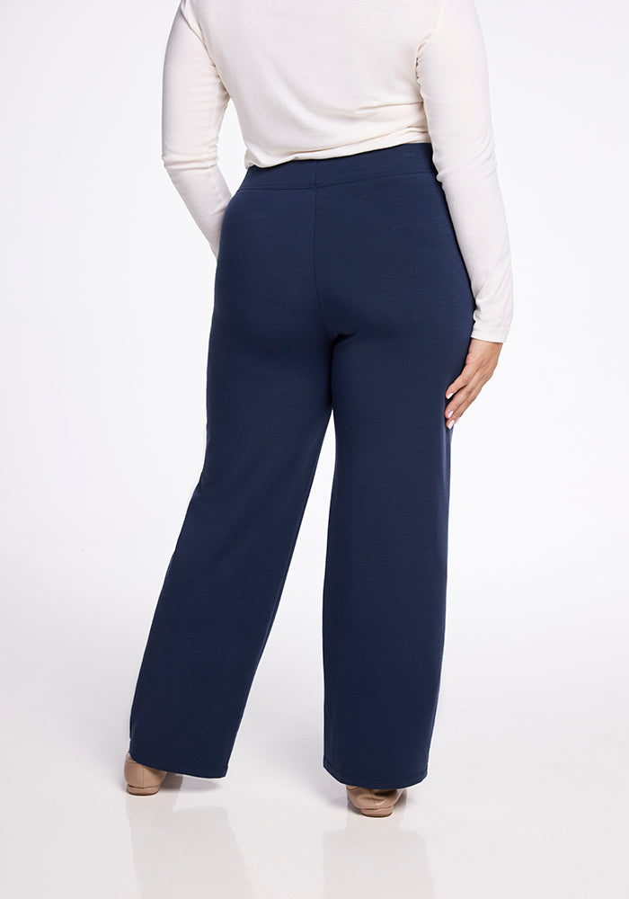 A rear view of a person wearing the Ellie Wide Leg Pants in Deep Navy by Woolx, paired with a white long-sleeve top. They are standing on a light-colored floor with beige shoes visible.