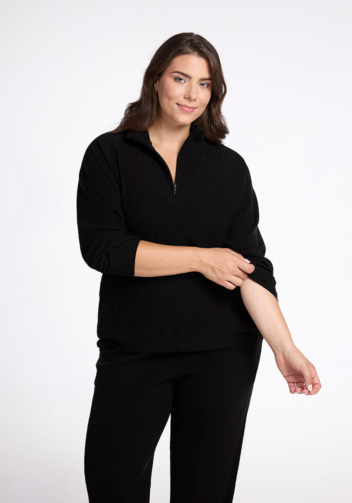 A person with long brown hair is smiling gently while wearing the Woolx Quinn Ribbed 1/4 Zip in black along with black pants. They are rolling up their left sleeve against a plain white background. 