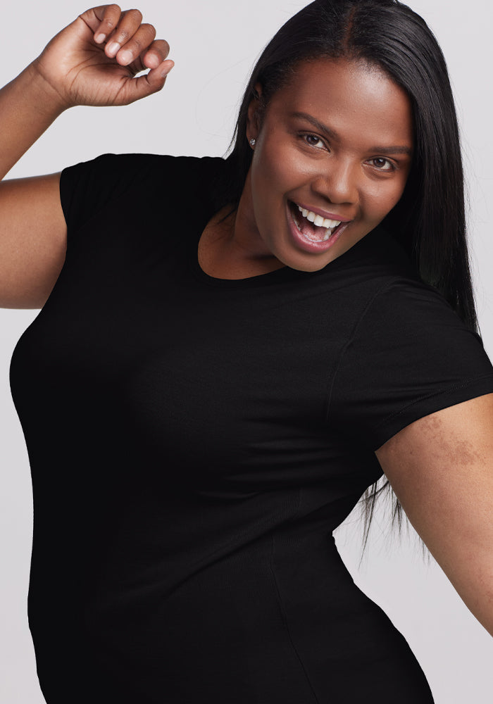 A joyful woman with long dark hair wears the ultra-soft Woolx Addie Crew Neck in black, posing with one arm raised and her mouth open against a light background.