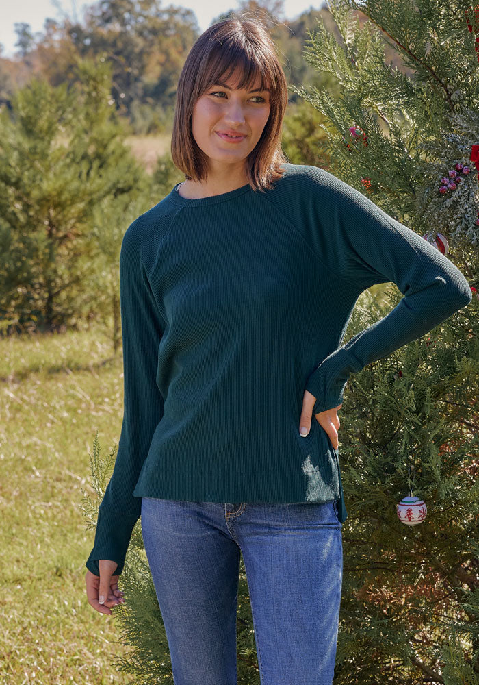 A woman with short brown hair wearing the Woolx Ainsley Sweater in black and blue jeans smiles while standing outdoors near a decorated evergreen tree.