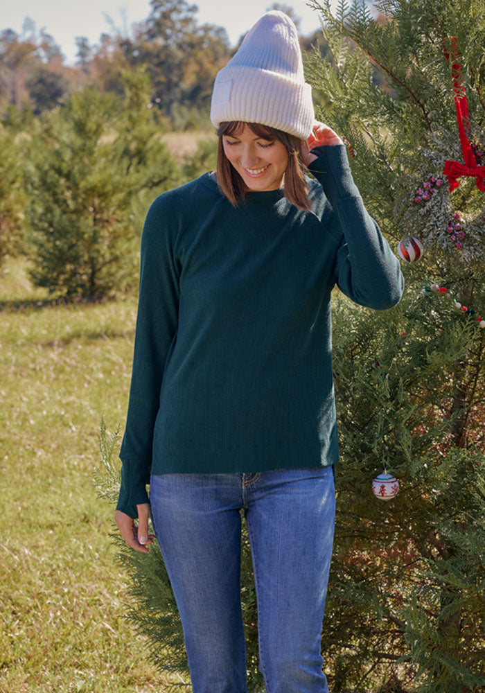 A woman wearing a cream beanie, blue jeans, and the Woolx Ainsley Sweater in black stands outdoors by a decorated Christmas tree, smiling and looking down. Green trees and grass fill the background.