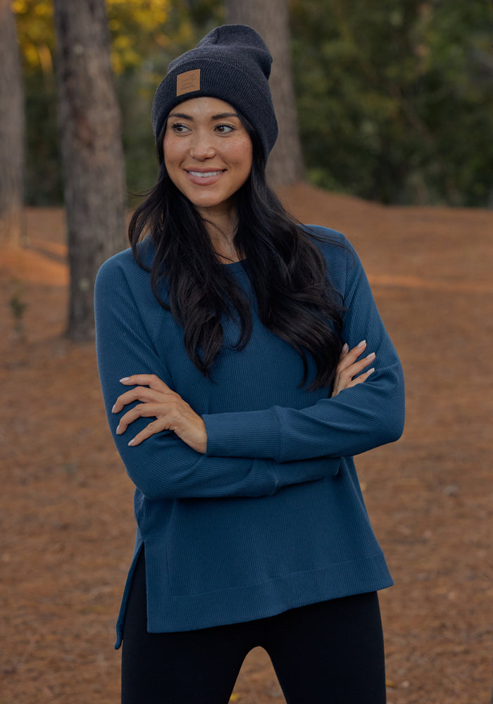 A woman with long dark hair, wearing a dark knit beanie, black pants, and a Woolx Ainsley Sweater in Black stands outdoors with her arms crossed, smiling in a wooded area with pine-covered ground.