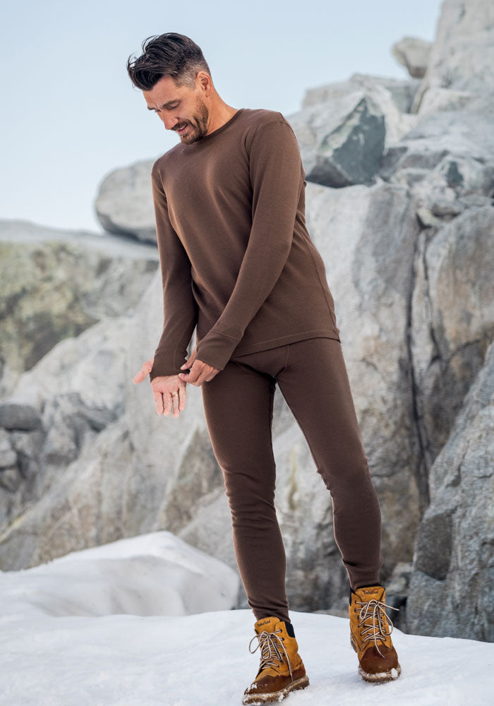 A man in Woolx Arctic Leggings and tan hiking boots stands on snow near large gray rocks, looking down and adjusting his sleeve.