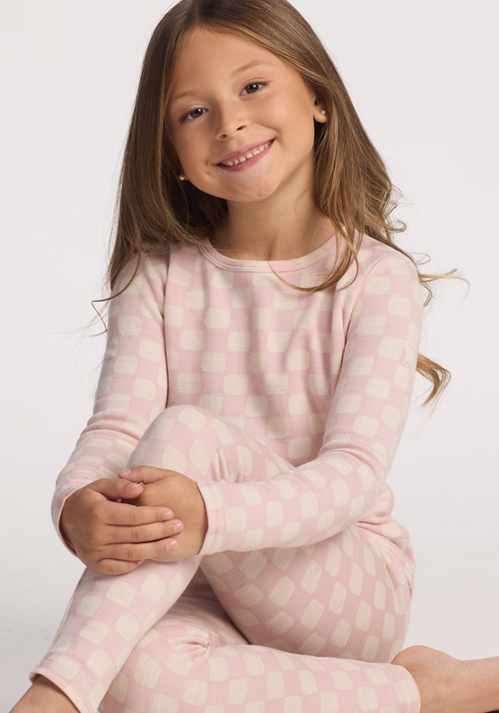 A young girl with long brown hair sits cross-legged and smiles at the camera, wearing the Woolx Kids Explorer Base Layer Top in Hopscotch Pink, made from soft Australian Merino wool, against a plain white background.