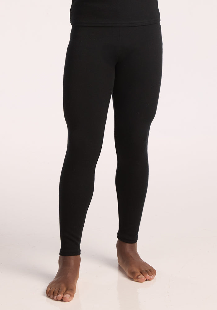 A person stands barefoot on a light surface, showing only the lower half of their body, while wearing Woolx Kids Explorer Base Layer Bottoms in black.