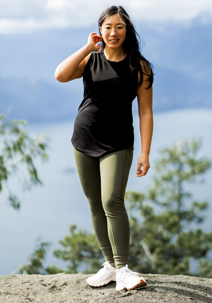 A woman smiles outdoors on rocks, wearing Woolx Merino Flex® McKenna Pocket Leggings in Everleaf, a black sleeveless top, and white sneakers. Blurred trees and a large body of water are in the background.