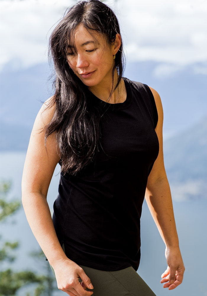 A woman with long dark hair, wearing the Woolx Bella Tunic Top in black—a true summer staple—stands outdoors, smiling slightly as she looks down. Blurred mountains and a lake can be seen in the background.