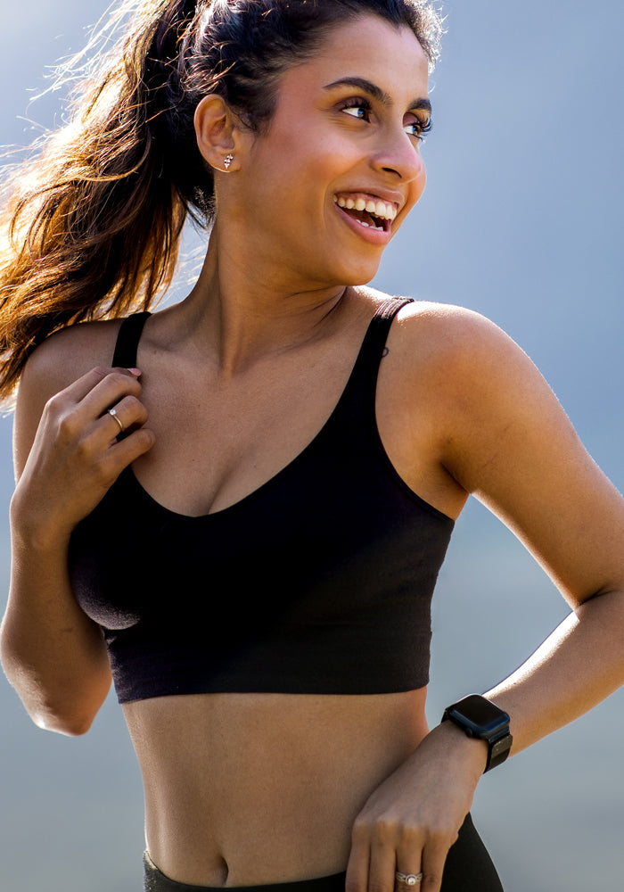 A woman with long brown hair in a ponytail smiles outdoors, wearing the black Woolx Hayden Bra with seamless construction, along with a fitness watch, earrings, and a ring. Sunlight highlights her face against a blurred blue backdrop.