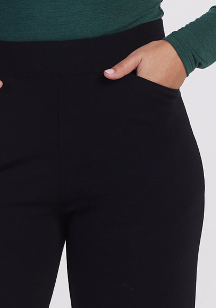 A petite woman is dressed in a stylish green long-sleeve top and Ellie Wide Leg Pants - Petite by Woolx, with one hand in her pocket. The emphasis is on her outfit, showcasing the fit and style of the wide-leg pants and chic top.