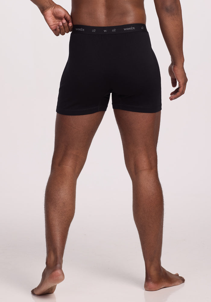 A person stands facing away, wearing Woolx's Breathe Boxer Shorts - Black. The subtly prominent branding label on the waistband complements the plain, well-lit background that highlights their breathable Woolmark® Certified Merino quality.