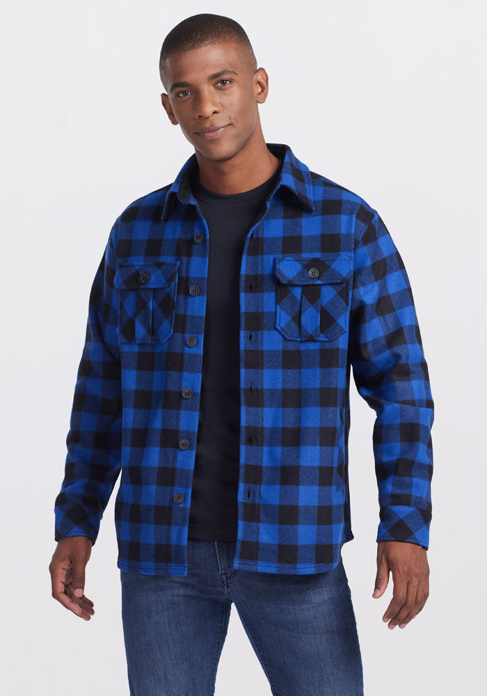A man wearing a Woolx Burlington Jacket in bright blue and black plaid over a moisture-wicking black t-shirt, paired with jeans. He stands against a plain white background, smiling slightly at the camera.