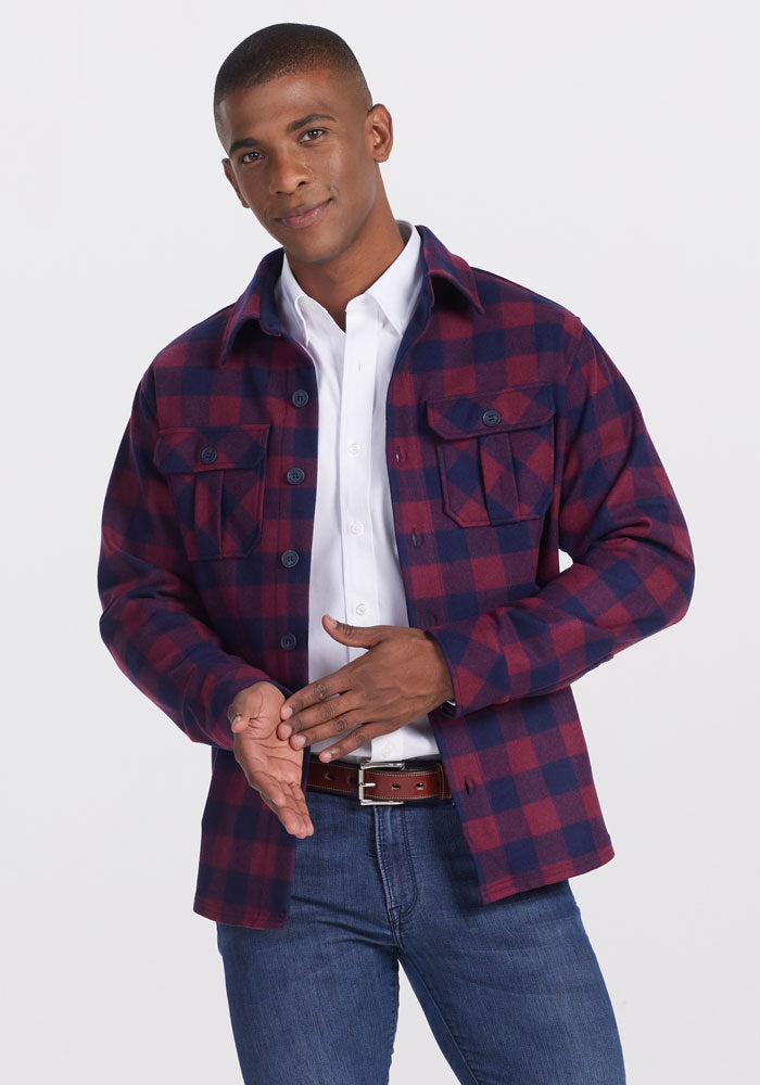 A man with short hair and a slight smile faces forward, wearing a white shirt, blue jeans, and the Woolx Burlington Jacket • Final Sale - Cranberry Navy Checkered, with one hand gesturing and the other on his belt.