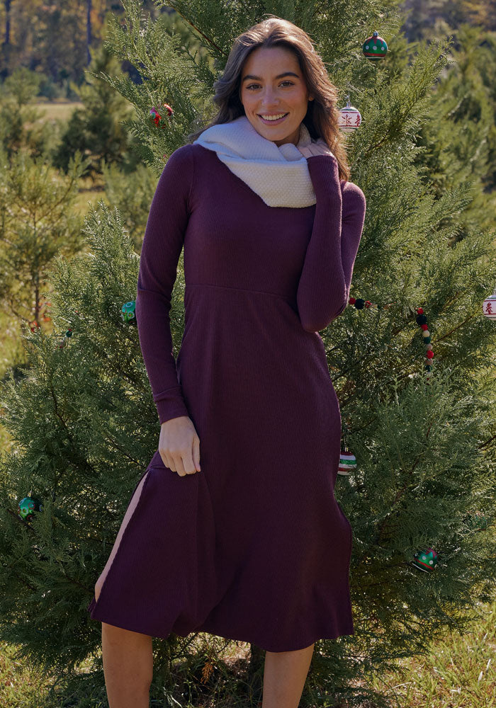 A woman smiles at the camera outdoors in front of a decorated evergreen tree, wearing the Woolx Caroline Ribbed Twirl Dress in Toasted Garnet with a light scarf, creating a festive winter holiday vibe.
