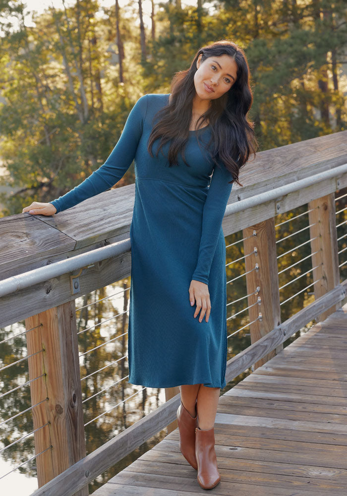 A woman with long dark hair wears the Woolx Caroline Ribbed Twirl Dress in Deep Timber and brown ankle boots as she stands on a wooden bridge, leaning on the railing with trees in the background.