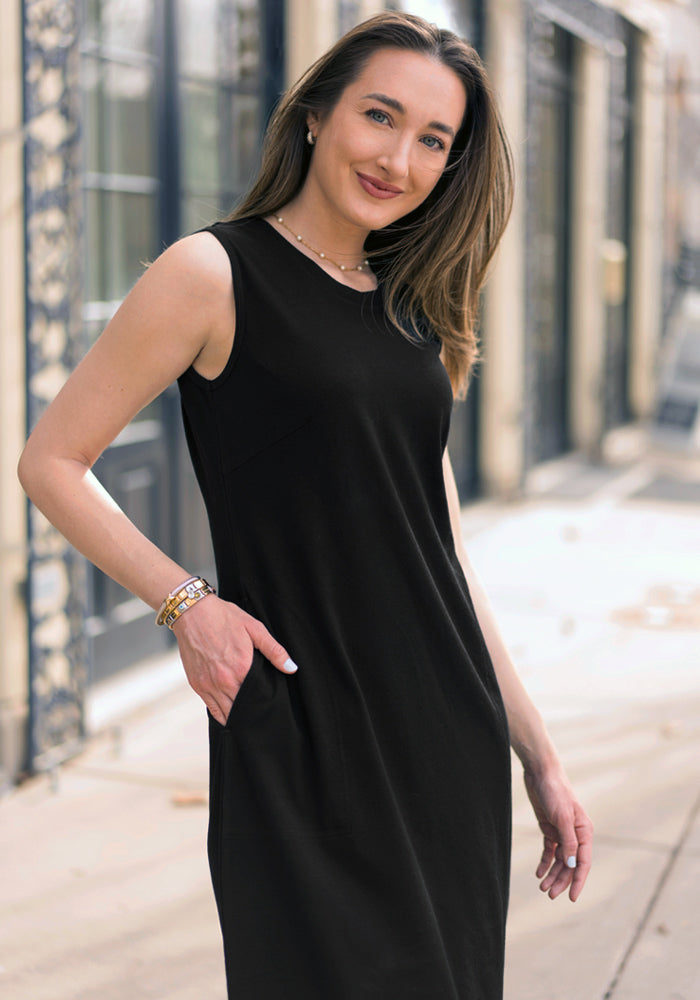 Woman in the Woolx Cassie Dress - Black, sleeveless Merino wool midi, stands outdoors smiling with a hand in her pocket.