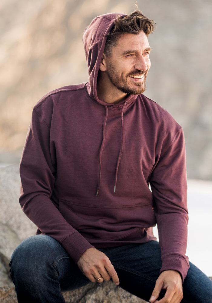 A man with short brown hair and a beard sits outdoors, smiling and looking to the side. He is wearing a Woolx Chase Hoodie in black with the hood up and dark jeans, set against a blurred natural background.