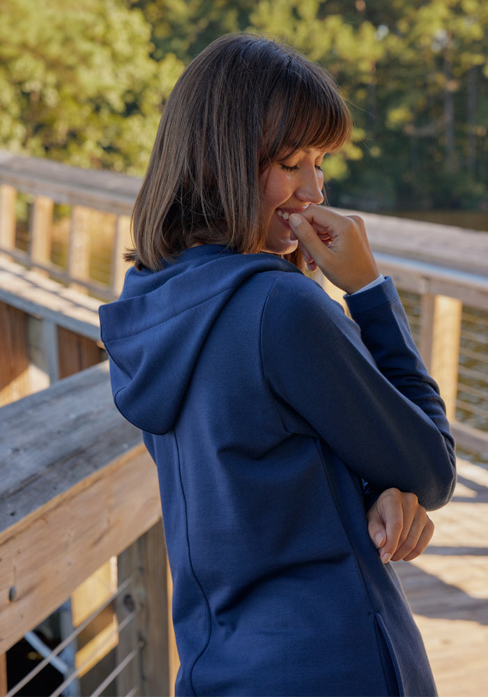 A woman wearing the Woolx Edna Hooded Cardigan in Cream Heather stands on a wooden boardwalk near water, smiling with her hand near her mouth. Trees and sunlight fill the background.