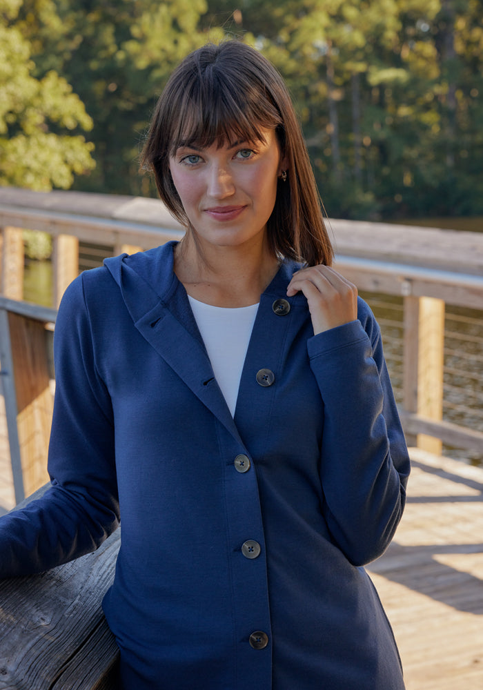 A woman with straight brown hair and bangs, wearing the Woolx Edna Hooded Cardigan in black over a white top, stands on a wooden bridge near water with trees behind her, looking at the camera and smiling softly.