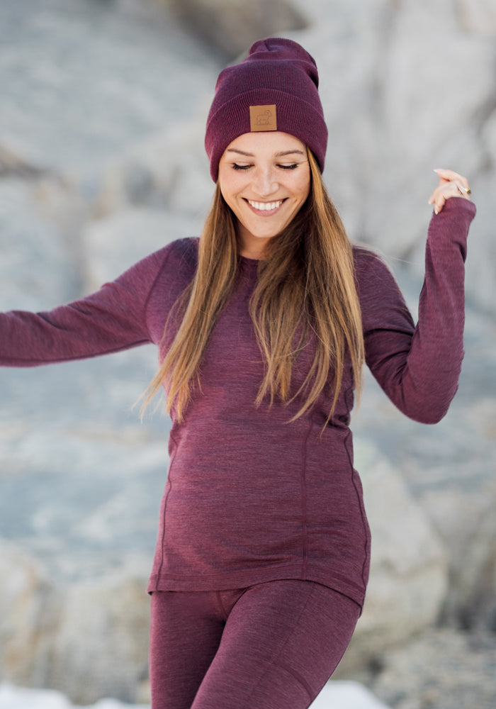 A smiling woman wears the Woolx Elsa Long Sleeve in black and a matching beanie, standing outdoors with blurred gray rocks in the background.