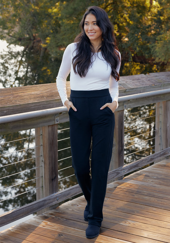 A woman with long dark hair, in a white long-sleeve top and Woolx Emerson Straight Leg Pants in Cinder Melange, stands smiling with hands in her pockets on a wooden boardwalk by the water amid trees.