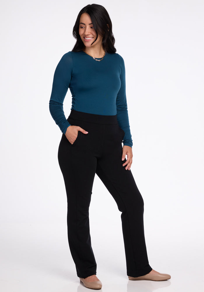 Dressed in a long-sleeve teal top and the stylish Emerson Straight Leg Pants in black by Woolx, a person stands with their left hand casually tucked into their pocket. They smile while glancing slightly to the side, set against a simple white background that enhances the sophistication of Merino Wool.