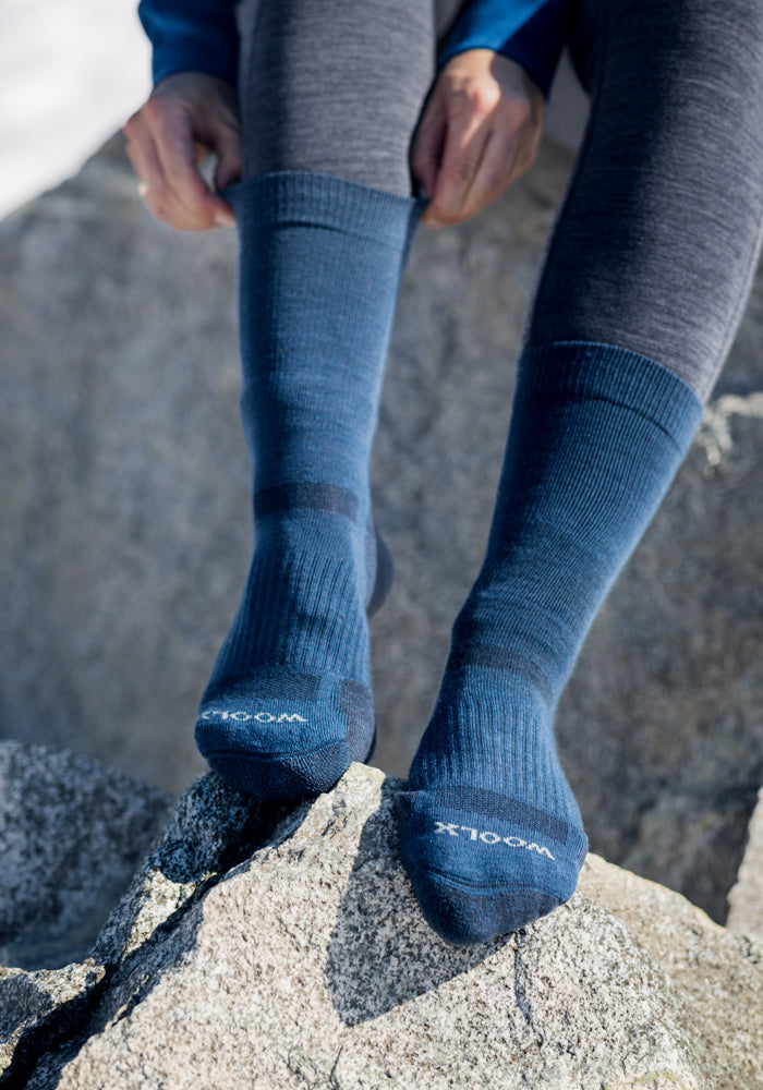 A person sits on a rock outdoors, adjusting their Womens Harbor Crew Sock in Majolica Blue by Woolx, worn over gray leggings. The dark blue Merino wool socks feature "WOOL" lettering on the toes.