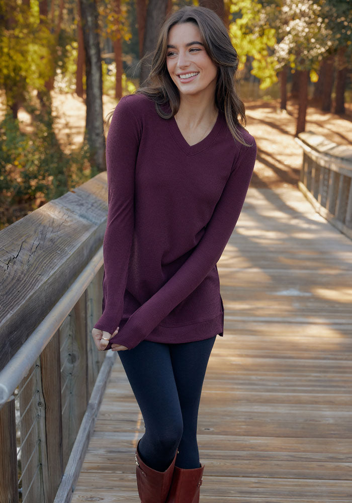 A woman in the Woolx Evie Ribbed Tunic - Black, paired with black leggings and brown boots, stands smiling on a wooden bridge in a forested area, looking to the side.