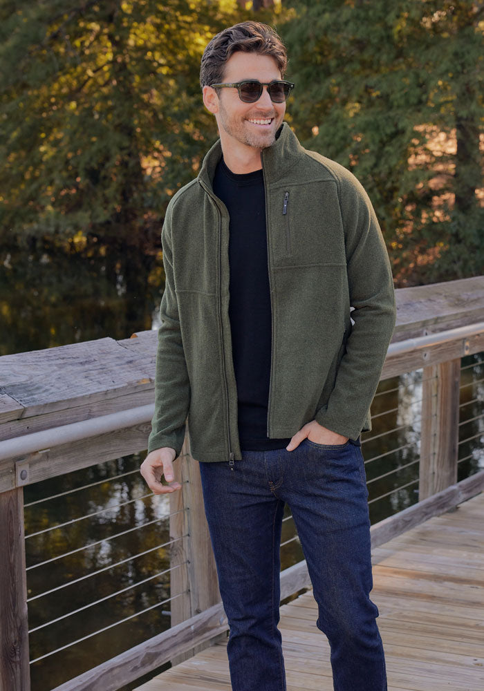 A man wearing sunglasses, jeans, and the Woolx Fairbanks Jacket in Carbon Black stands smiling on a wooden bridge with trees and water in the background.