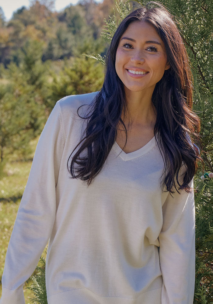 A woman with long dark hair smiles outdoors in sunlight, wearing the Woolx Francesca Sweater in Wild Salmon.