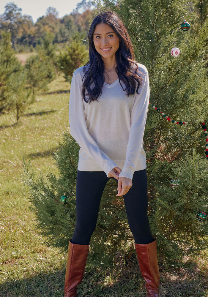 A woman with long dark hair, wearing the Woolx Francesca Sweater in black, black leggings, and brown boots, stands smiling in front of a decorated evergreen tree outdoors on a sunny day.