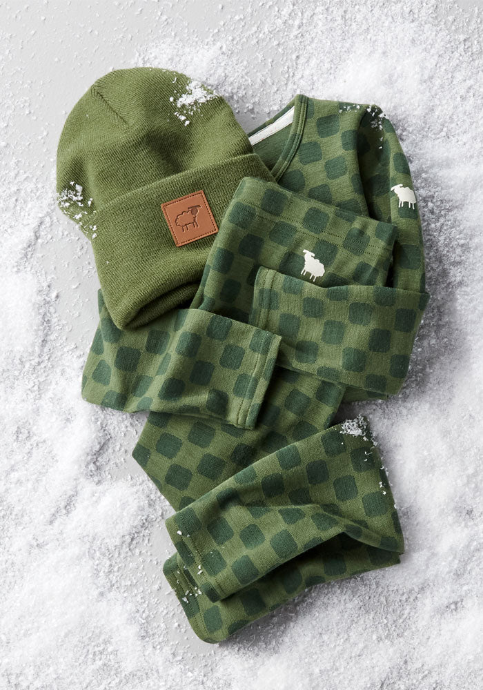A matching green checkered pajama set with a sheep logo, Woolx Kids Explorer Base Layer Bottoms in Frog Hop, and a green knit beanie with a brown patch are laid out on snow-dusted ground, with scattered snow patches on the clothing.