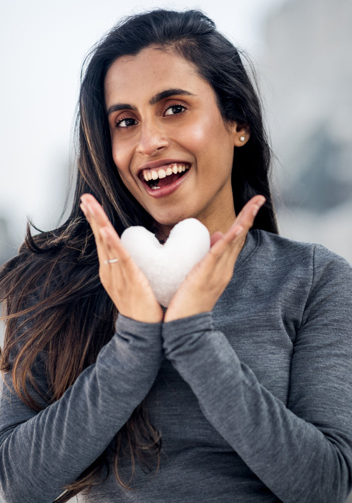 A smiling woman holds a heart-shaped snowball, wearing the Woolx Sadie Baselayer Top in Cinder Melange, made from heavyweight merino wool, with her long dark hair and a blurred outdoor background.