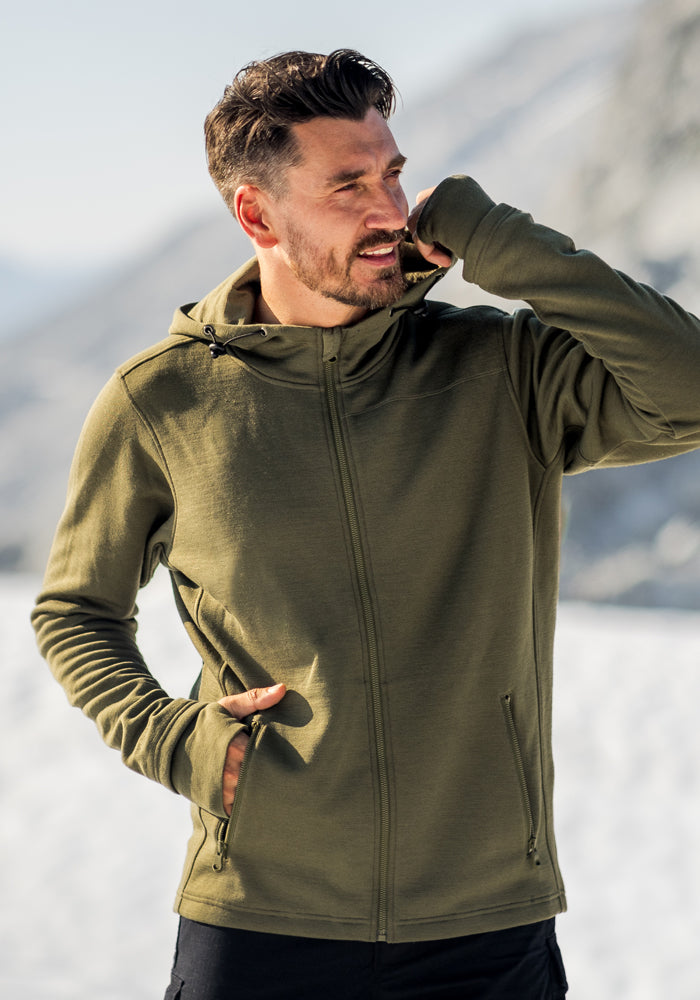 A man wearing the Woolx Grizzly Sweatshirt in Winter Moss stands outdoors in a snowy mountain landscape, with one hand near his mouth and the other in his pocket, looking to the side.